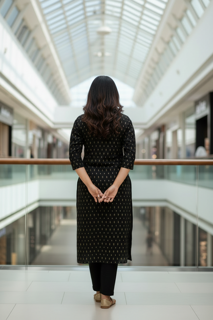 A woman stands in a modern shopping mall corridor wearing a black three-quarter sleeve kurta with a small white geometric pattern, paired with black pants and brown sandals.